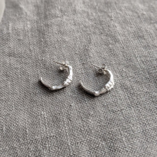 Recycled sterling silver midi hoop earrings with an organic surface by Lunaflux on a grey linen background.