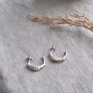 Recycled sterling silver midi hoop earrings with an organic surface by Lunaflux on a grey linen background.
