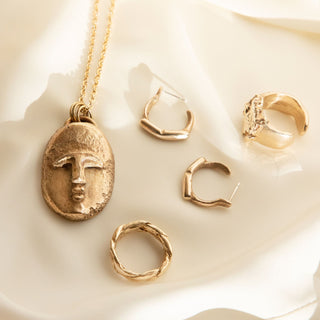 Recycled bronze face pendant with recycled gold rings and hoops on a gold silk surface.